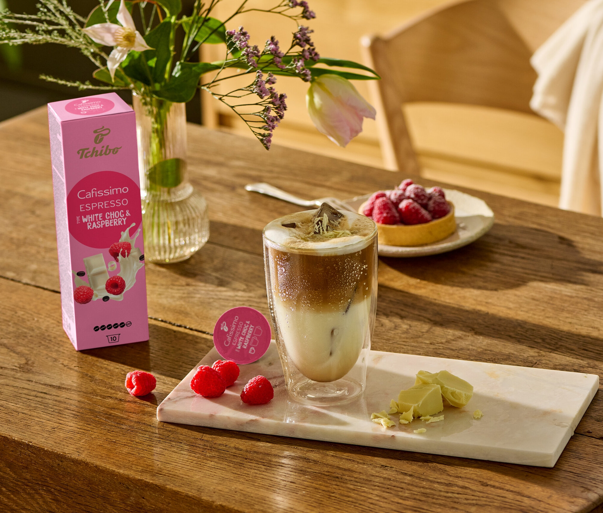 Egy fa asztalon egy pohár kávé, fehér csokoládé darabok és málna található egy márvány deszkán. A háttérben egy Cafissimo Flavoured Espresso - White Choc & Raspberry - 10 kávékapszula csomag látható.
