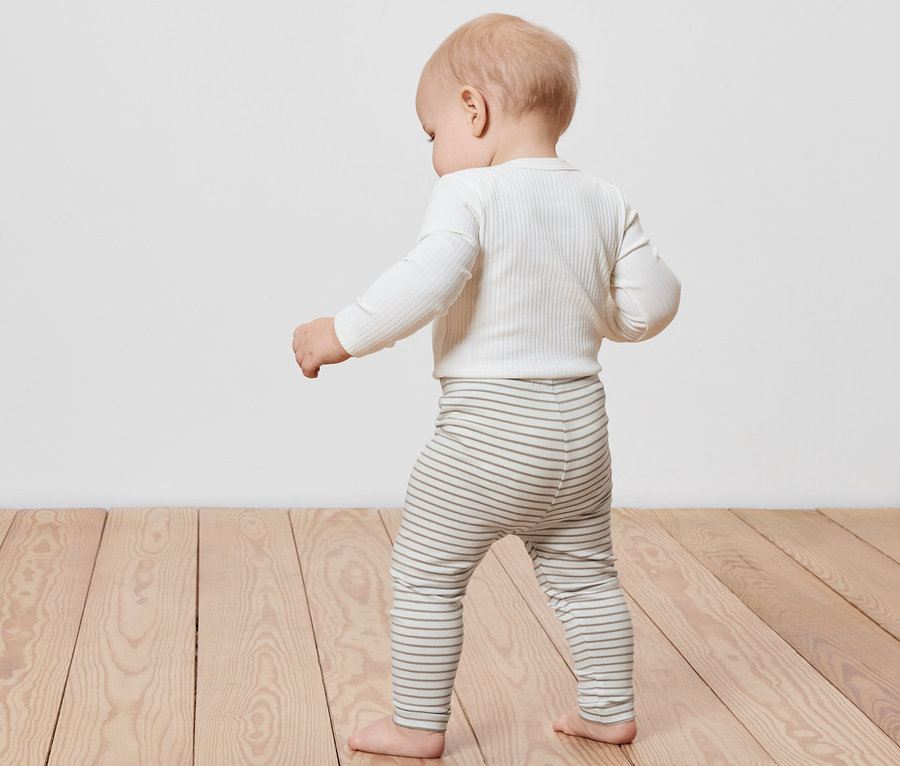 Kisgyermek sétál a fapadlón, csíkos 5 darabos gyerekleggingset visel.