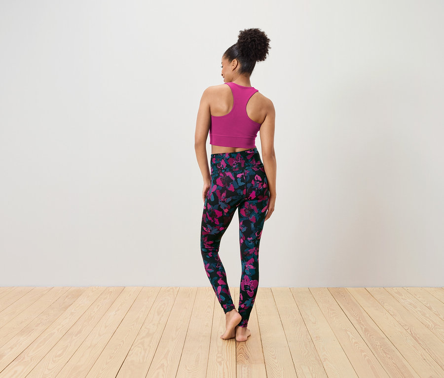 A fényképezőgépnek háttal álló nő rózsaszín sportmelltartót és rózsaszín-kék virágmintás fekete leggingset visel.