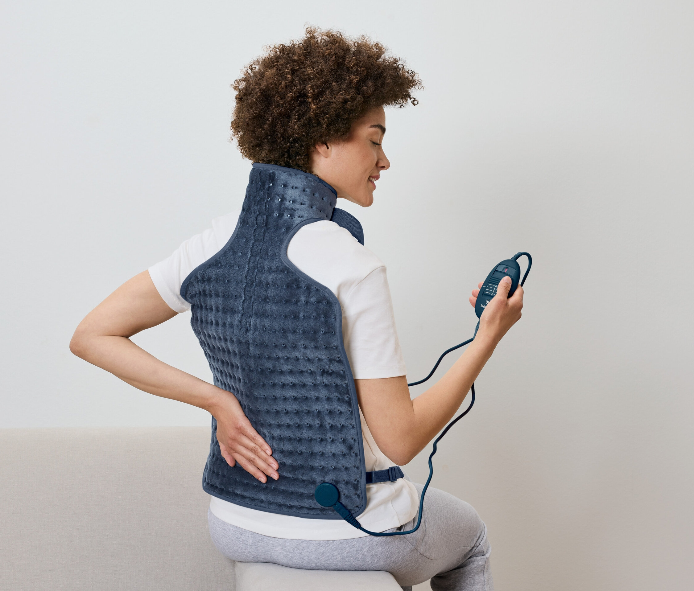 Egy nő ül és egy fűtött hátpárnát használ távirányítóval a kezében.