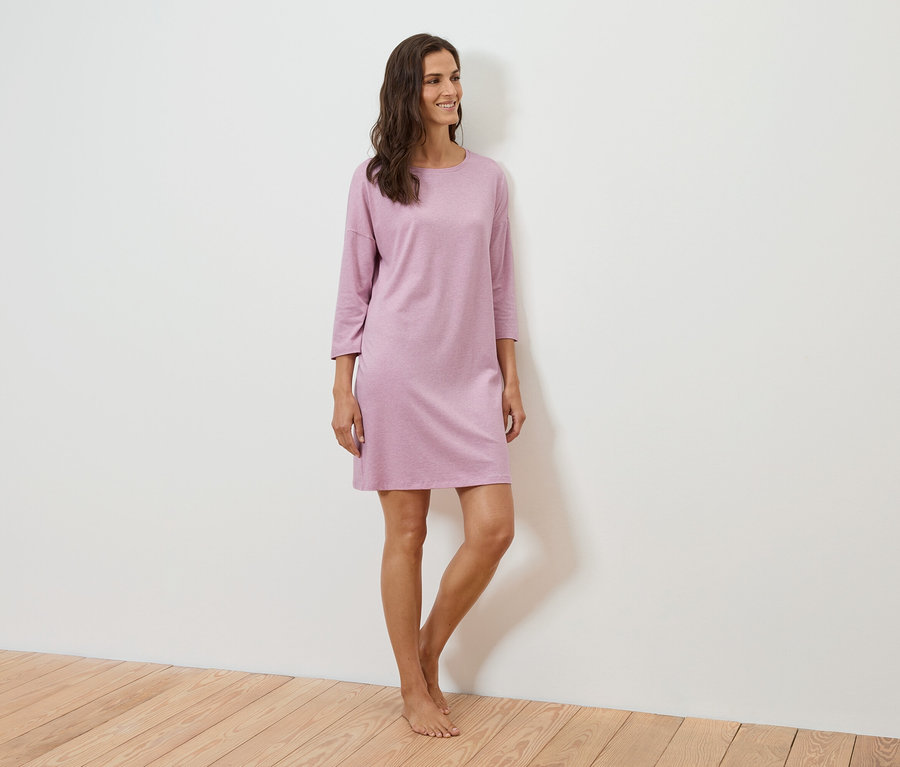 Egy nő áll és mosolyog, háromnegyedes ujjú lila ruhát visel.