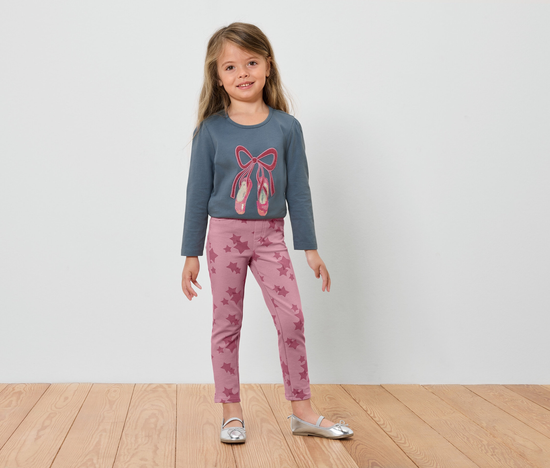 Kislány kék, balettcipős hosszú ujjúban és rózsaszín leggingsben áll a fapadlón.