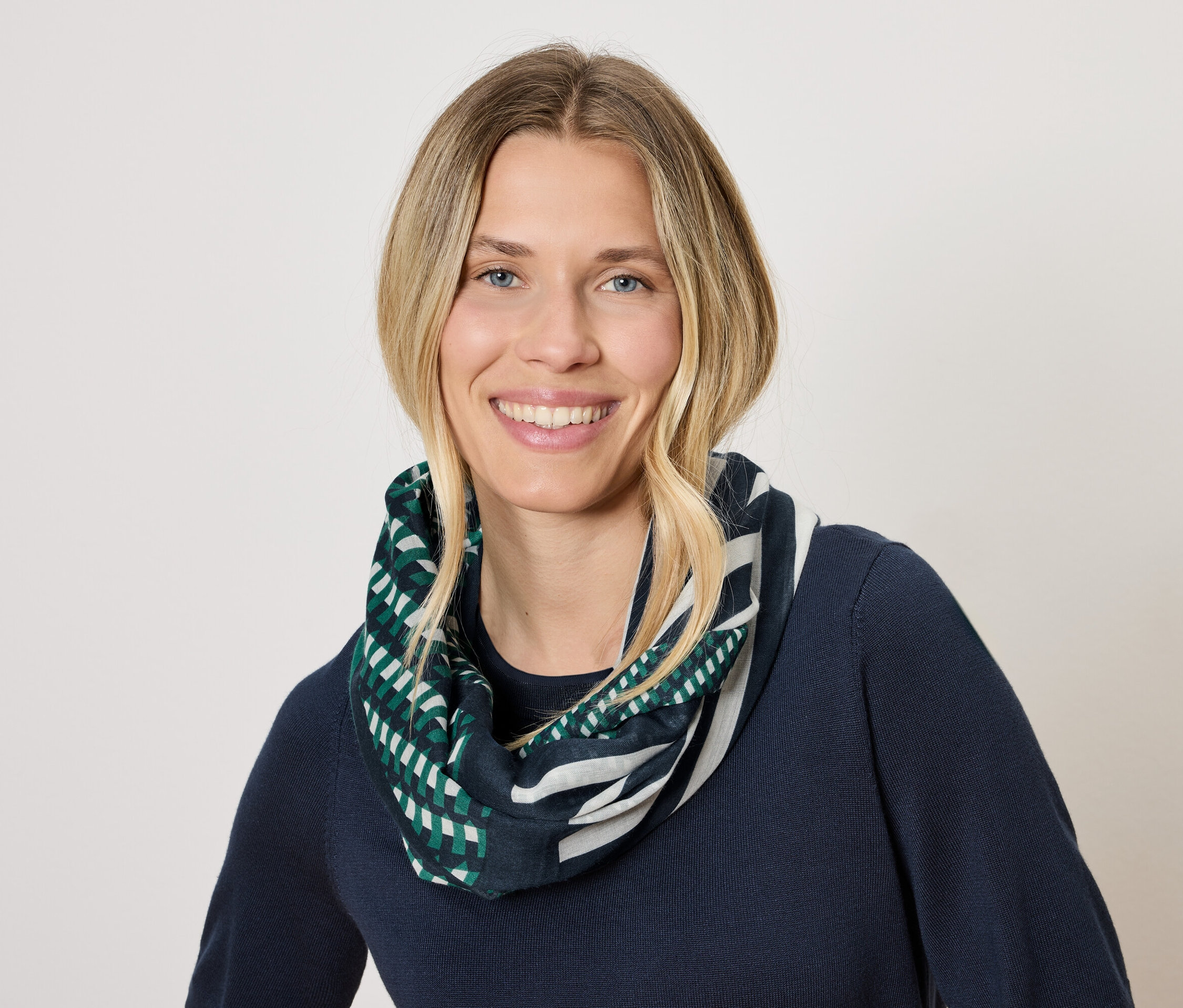 Egy szőke hajú, mosolygó nő sötétkék pulóvert és zöld-fehér geometrikus mintás sálat visel.