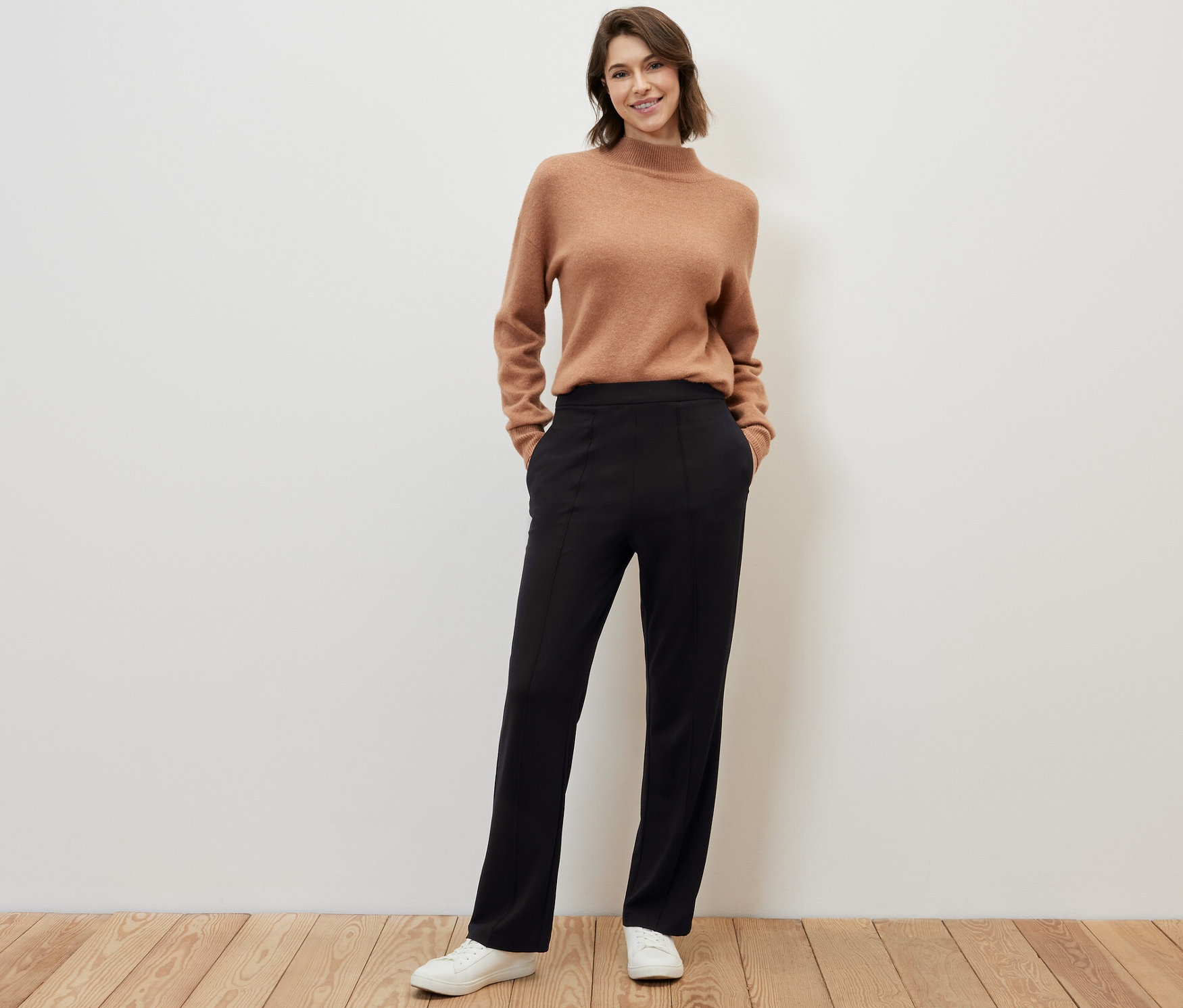 Nő pózol fapadlón barna pulóverben és fekete, levarrt élű jersey nadrágban.