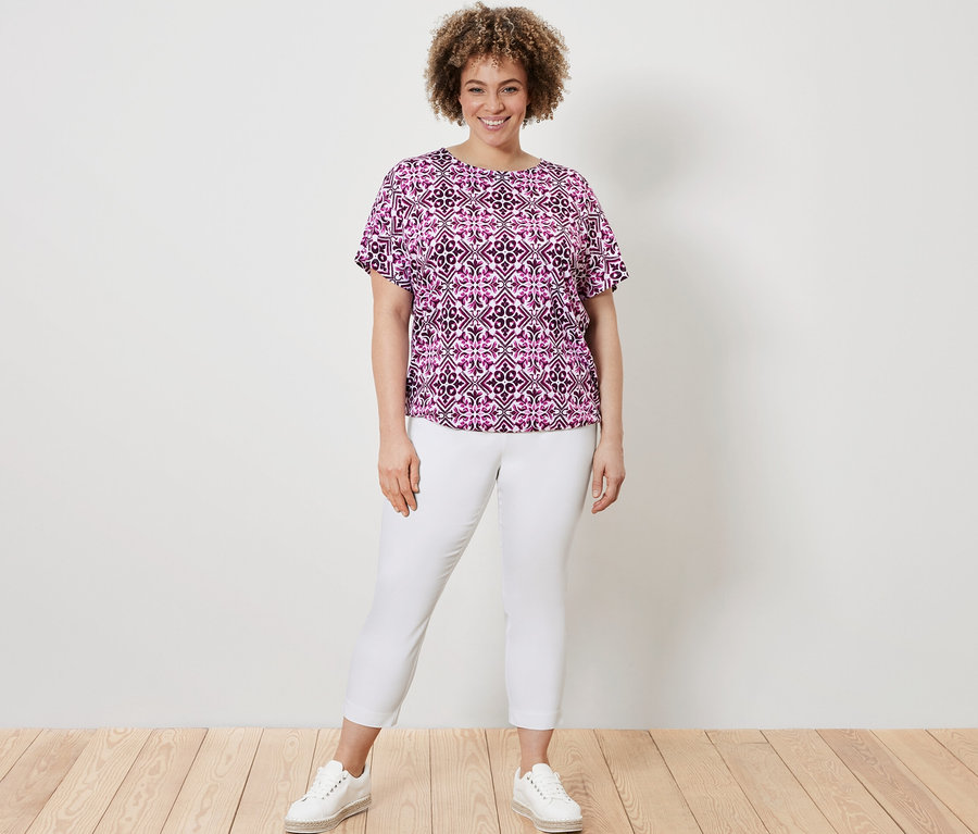 Nő pózol egy mintás fukszia női pólóblúzban és egy fehér női 7/8-os sztreccsnadrágban.