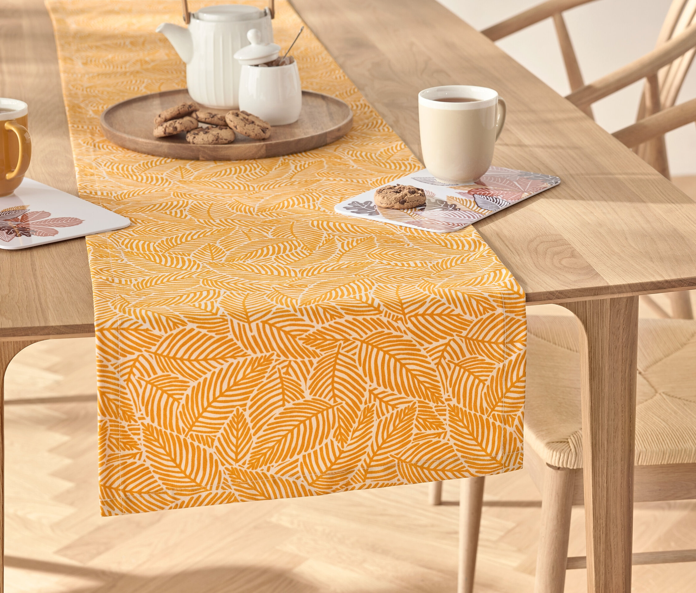 Egy fa asztalon egy sárga, leveles mintázatú asztali futó található, rajta egy teáskészlet, egy csésze és kekszek.