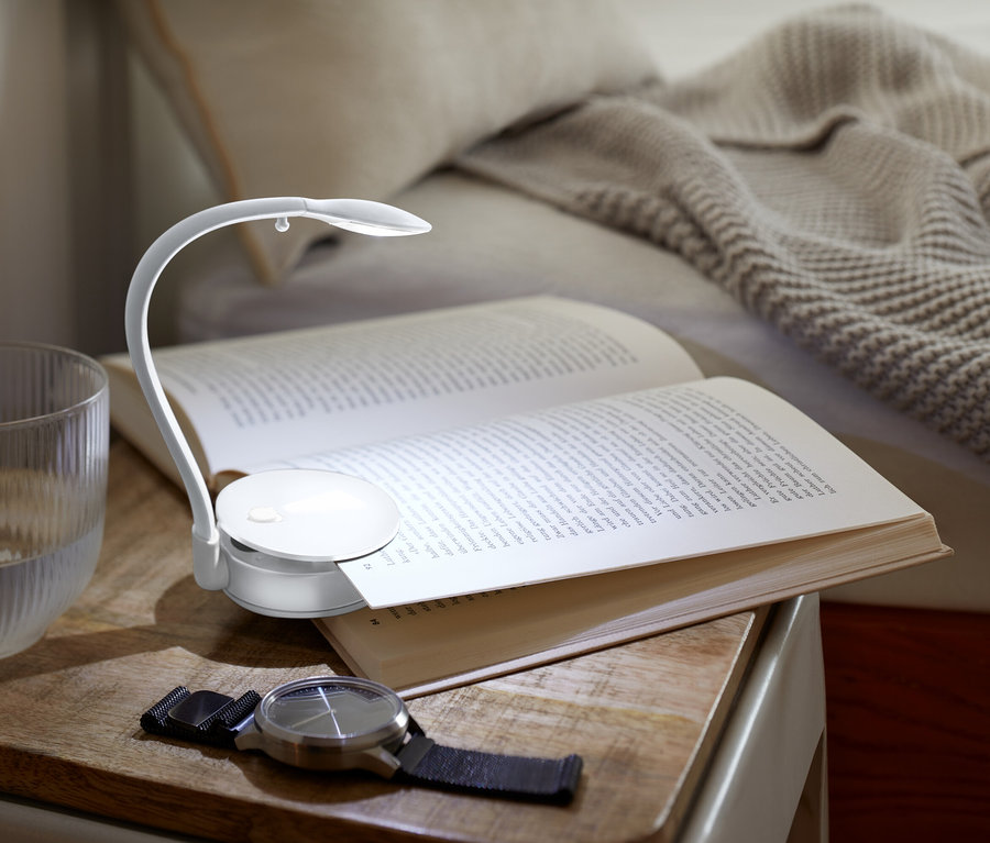 Az éjjeliszekrényen egy kinyitott könyv fekszik, amelyet egy kis lámpa világít meg, mellette egy pohár víz és egy óra.