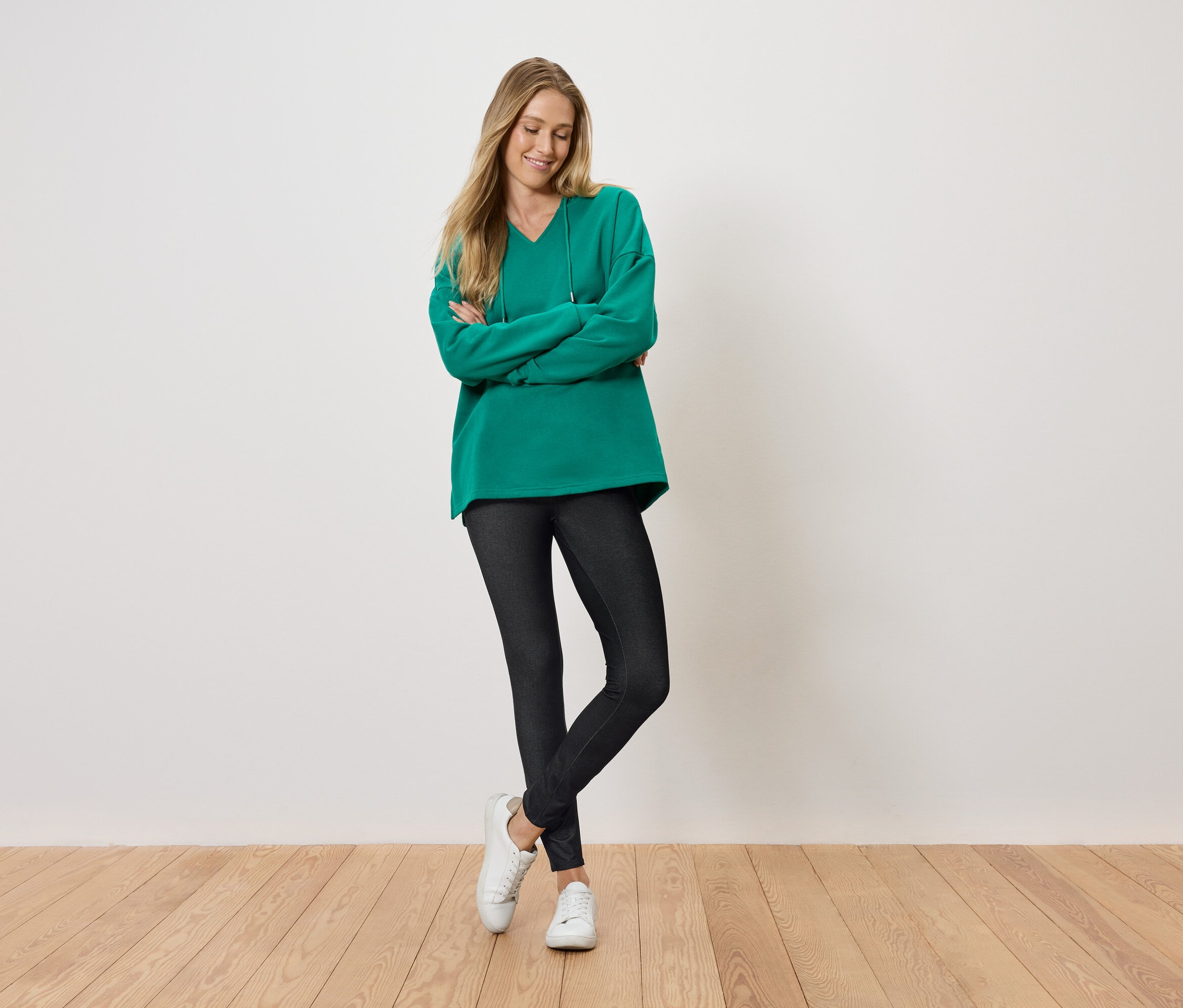 Egy nő áll keresztbe tett karokkal és mosolyog, zöld pulóvert, fekete leggingset és fehér cipőt visel.