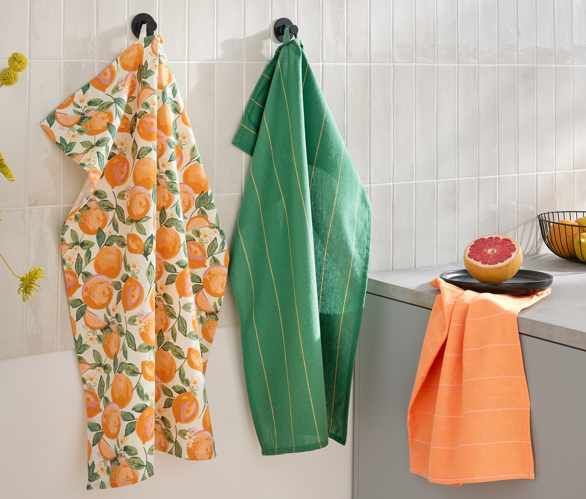 Három konyharuha lóg a falon: egy citrusmintás, egy zöld csíkos és egy narancssárga. A pulton egy félbevágott grapefruit és egy gyümölcsöstál található.