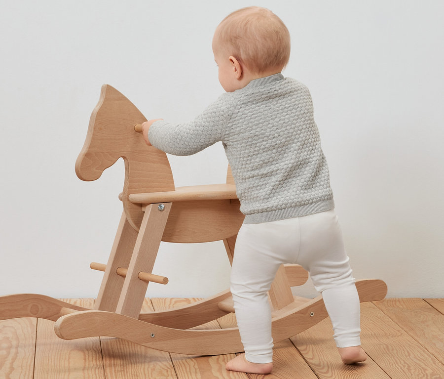 Kisgyermek kötött kardigánban egy fa hintalóhoz dőlve.