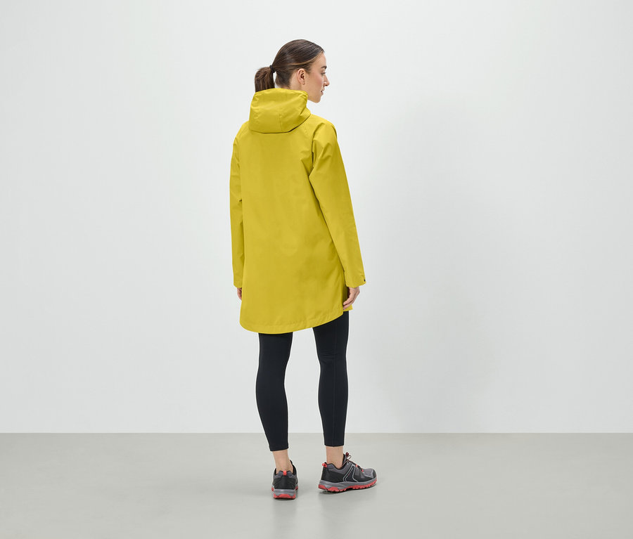 Egy nő sárga kapucnis kabátban, fekete leggingsben és sportcipőben áll háttal a kamerának.