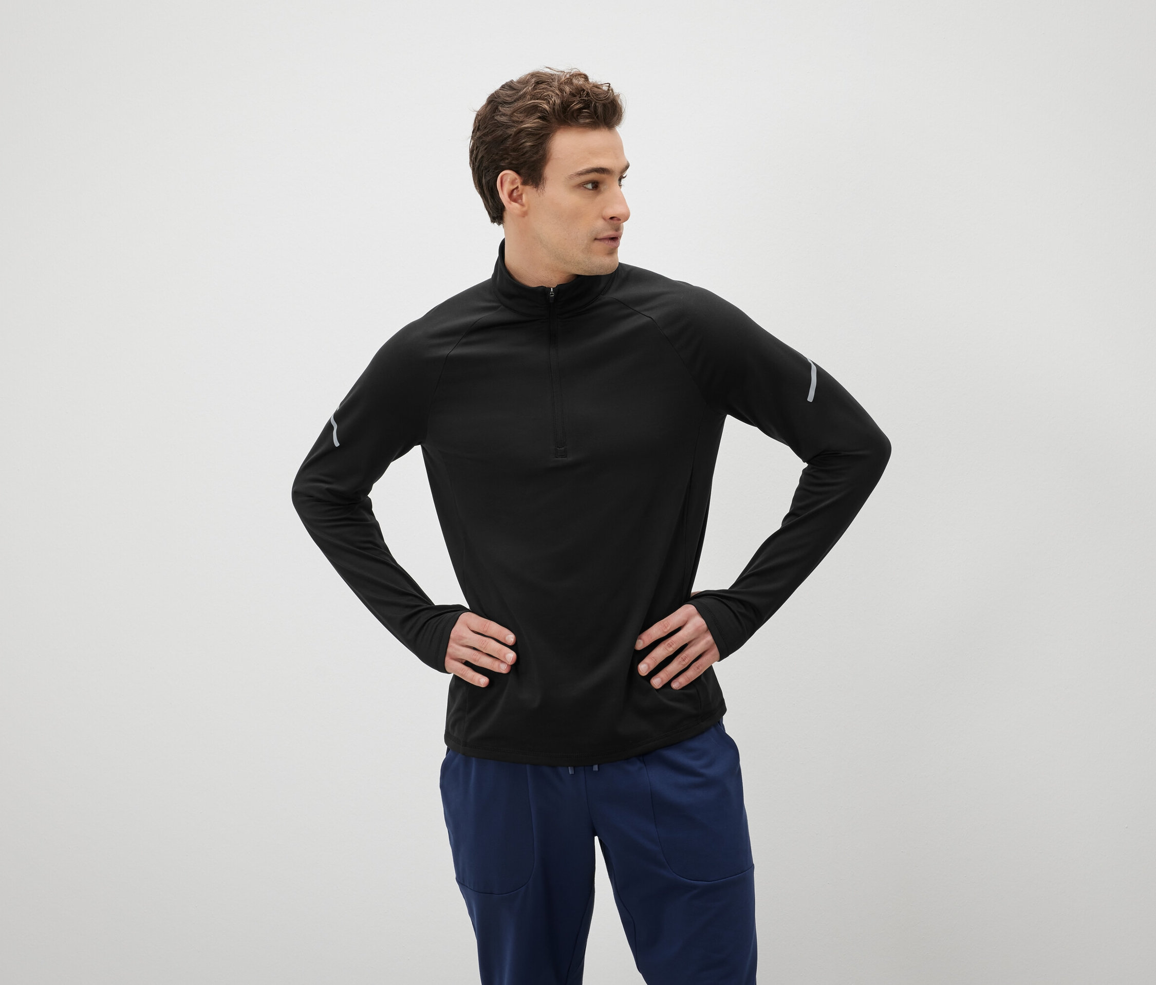 Barna hajú férfi pózol csípőre tett kézzel, fekete hosszú ujjú pólót és kék nadrágot visel.