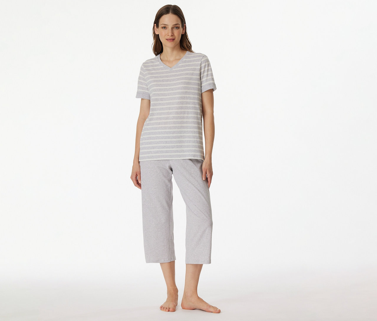 Nő visel SCHIESSER 3/4-es pizsamát szürke-fehér csíkos pólóval és szürke 3/4-es nadrággal fehér háttér előtt.