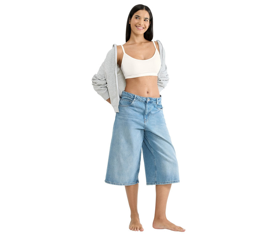 Egy nő áll mezítláb, fehér crop topot, bő farmer rövidnadrágot és szürke kabátot visel.