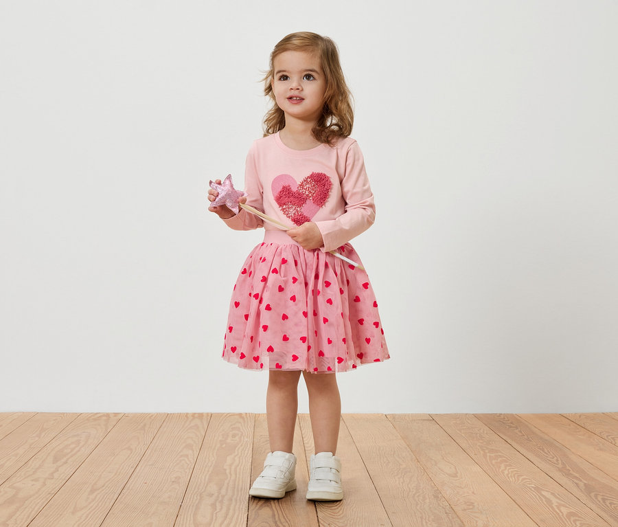 Egy szőke kislány rózsaszín, szívecskés ruhát visel, és egy varázspálcát tart.