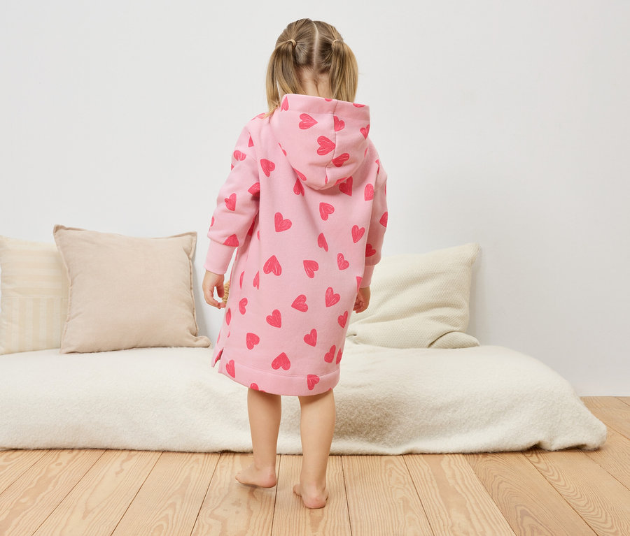 Egy kislány háttal áll a kamerának, rózsaszín, szívecskés, kapucnis ruhát visel.