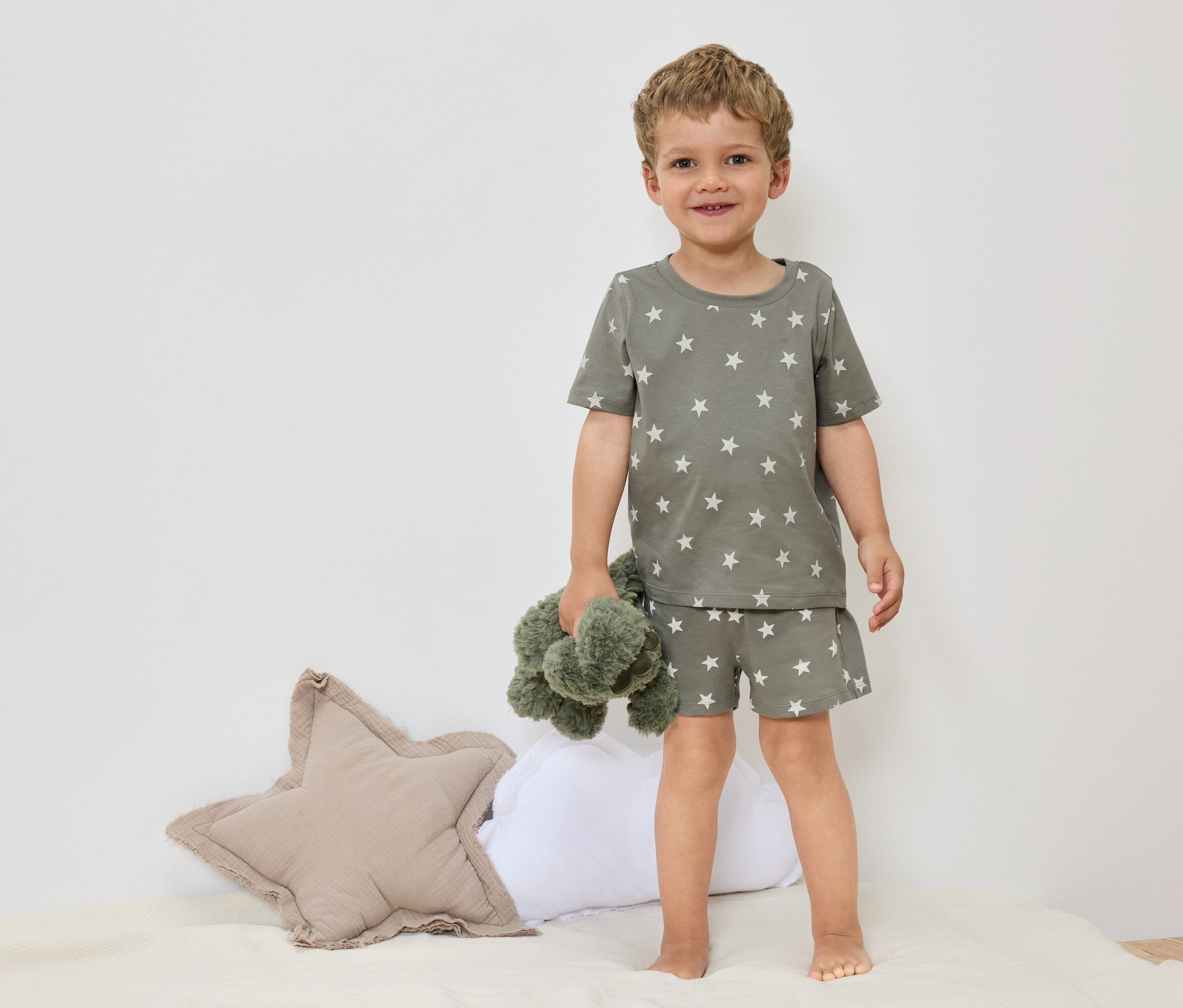 Egy kisfiú mezítláb áll az ágyon, olívazöld, csillagos pizsamában, és egy zöld plüssállatot tart a kezében.