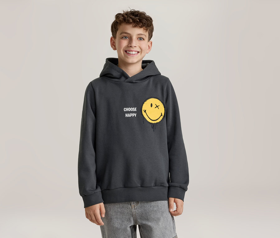 Mosolygó fiú sötétszürke kapucnis pulóverben, amelyen sárga, X szemű, olvadó hatású smiley arc és „CHOOSE HAPPY” felirat látható. Világosszürke farmert visel.