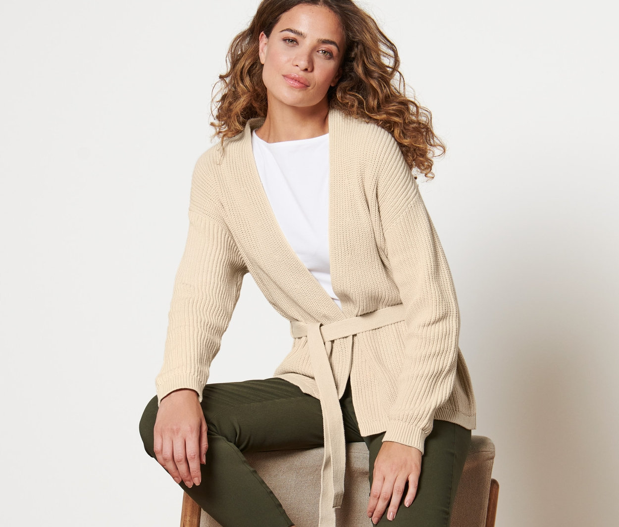 Nő ül egy széken, és bézs színű, átlapolós kardigánt visel.