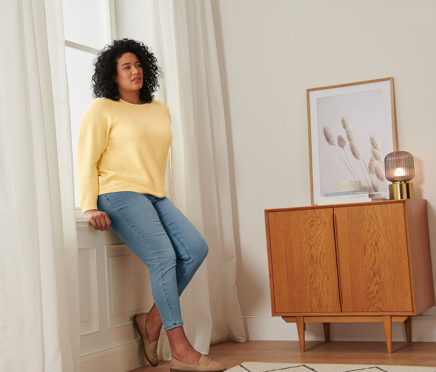 Nő ül az ablakpárkányon kötött, sárga pulóverben, treggingsben és velúrbőr mokaszinban.