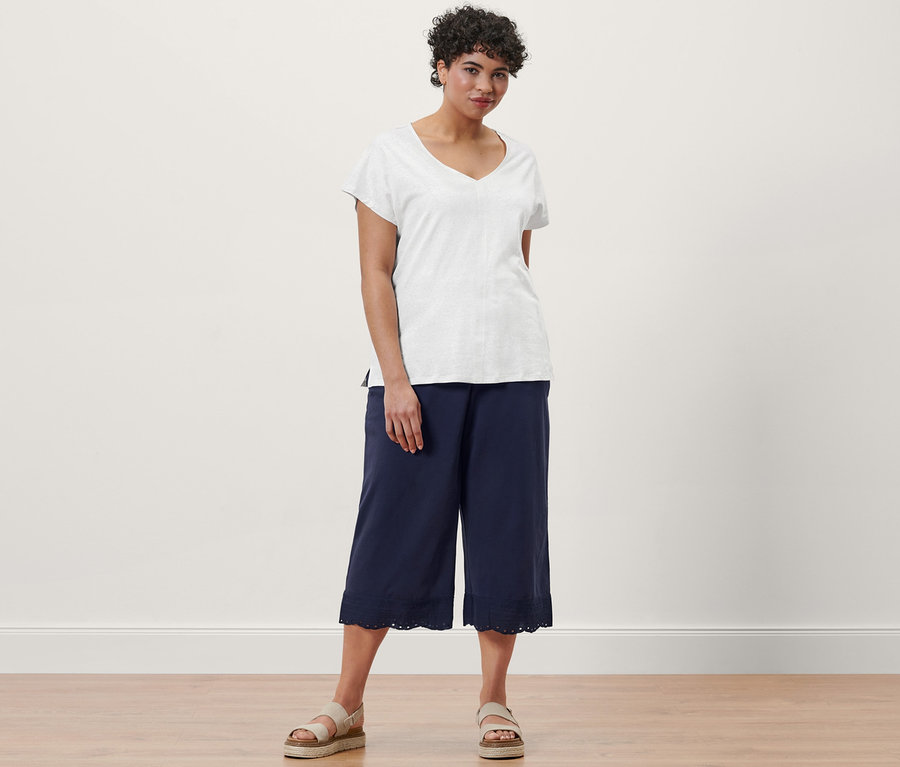 Nő pózol krémfehér len keverékéből készült pólóban, sötétkék, hímzett culotte nadrágban és bőr platform szandálban.