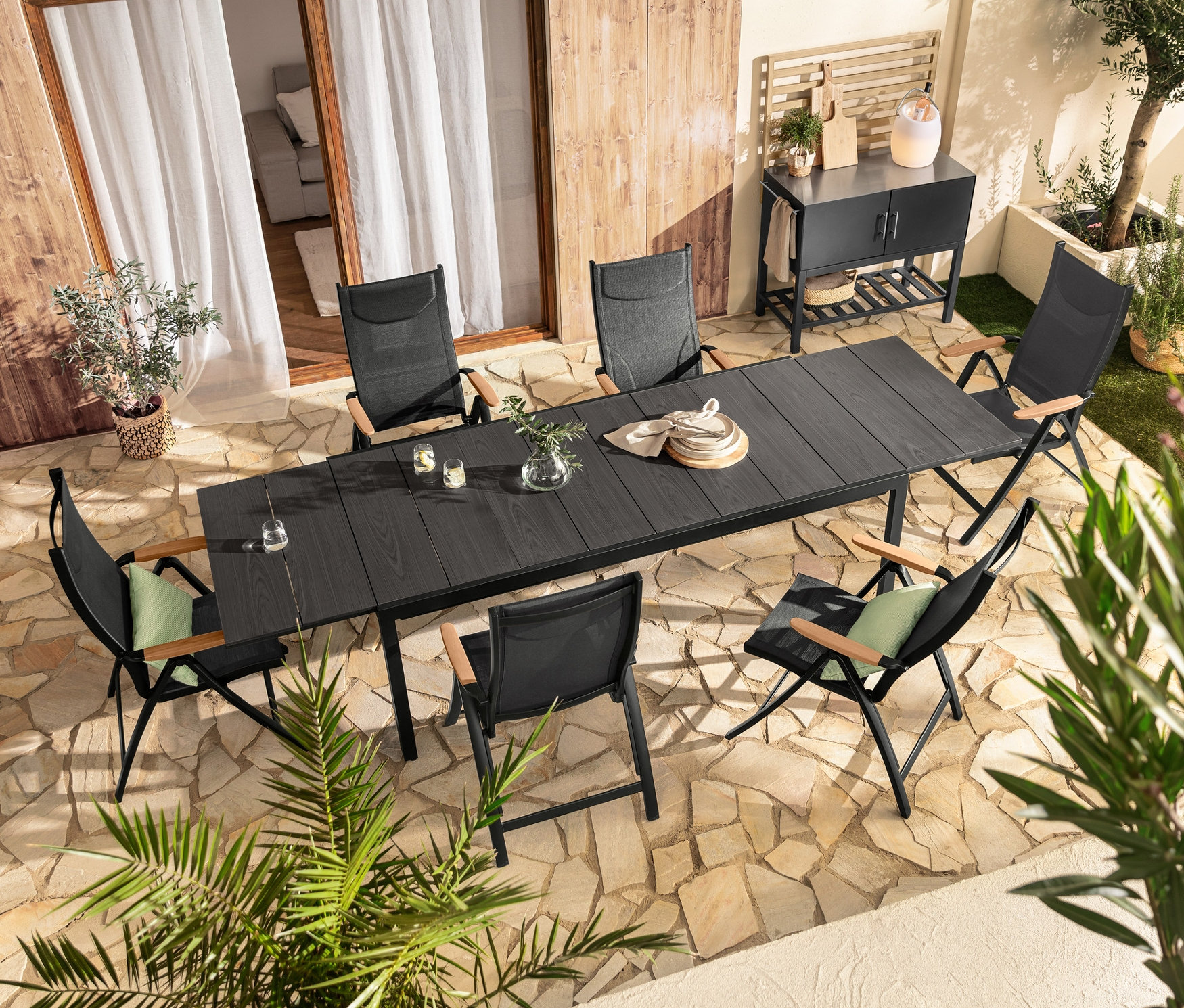 Egy fekete, Duraboard® asztallappal ellátott kihúzható asztalt magas háttámlás székek vesznek körül, és a közelben 2 »Tinus« virágtartó láda áll.