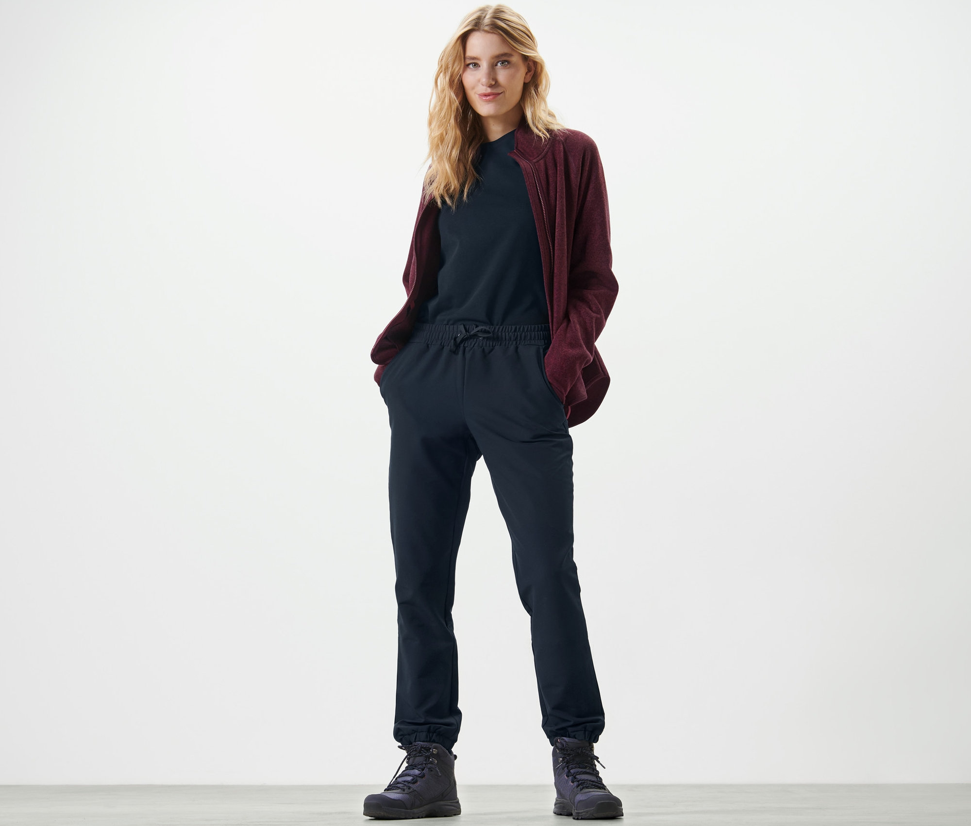 Egy szőke nő áll, kezével a sötétkék softshell lezser nadrág zsebében.