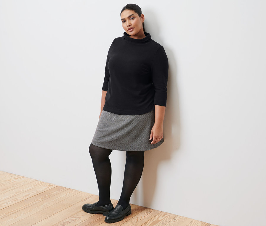 Egy nő a falnak dőlve fekete, 3/4-es ujjú szabadidőfelsőt, szabadidőszoknyát, fekete harisnyát és bőr mokaszint visel.