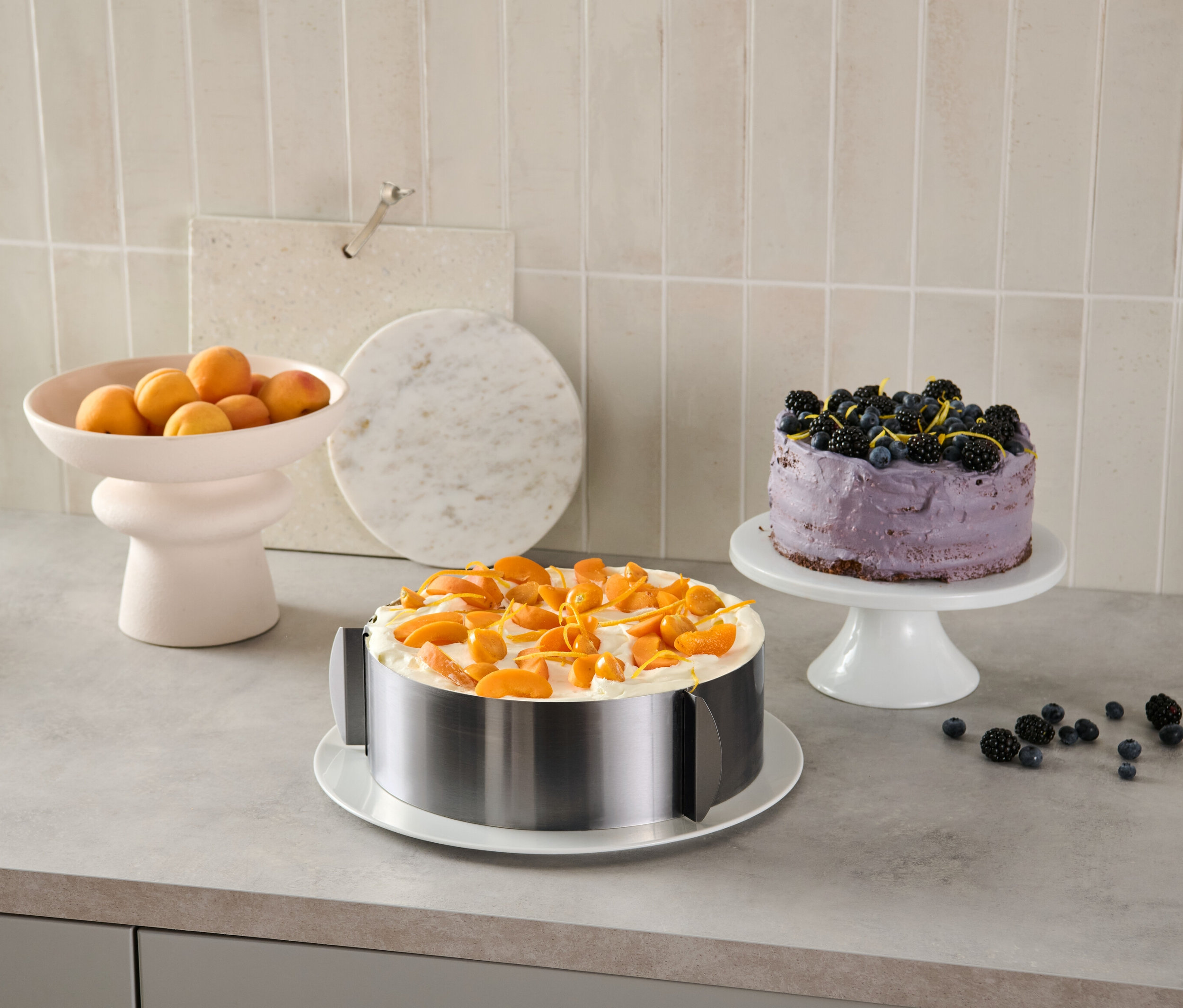 Egy sárgabarackkal díszített torta egy nemesacél tortagyűrűben van. A háttérben egy áfonyás torta és egy tál sárgabarack látható.