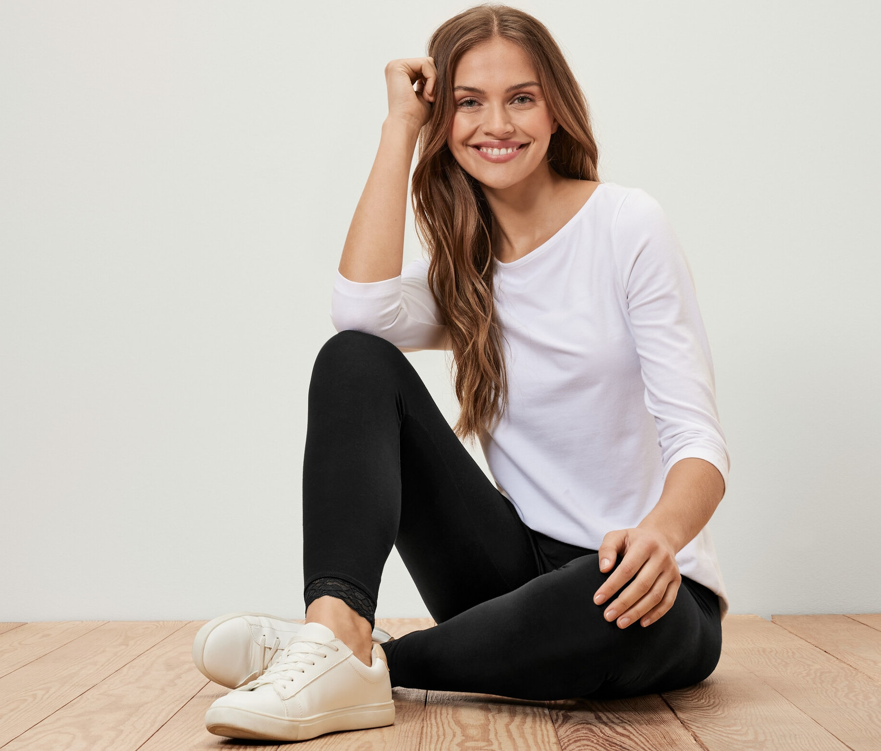 Egy nő ül egy fapadlón, fekete, csipkeszegélyes leggingset, fehér, háromnegyedes ujjú pólót és fehér tornacipőt visel.