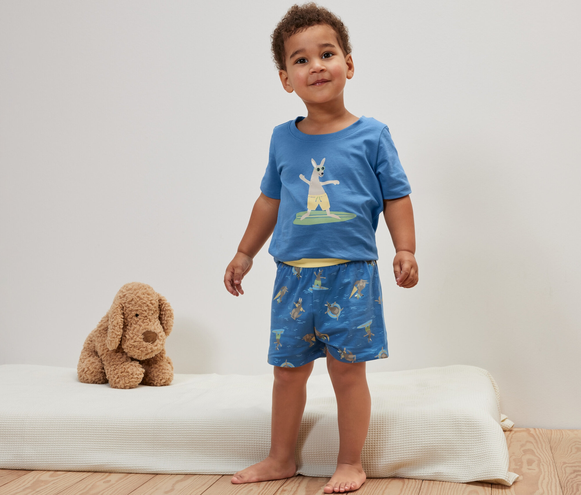 Egy kisfiú áll egy matracon, kék, rövidnadrágos gyerekpizsamában, kenguru mintával.