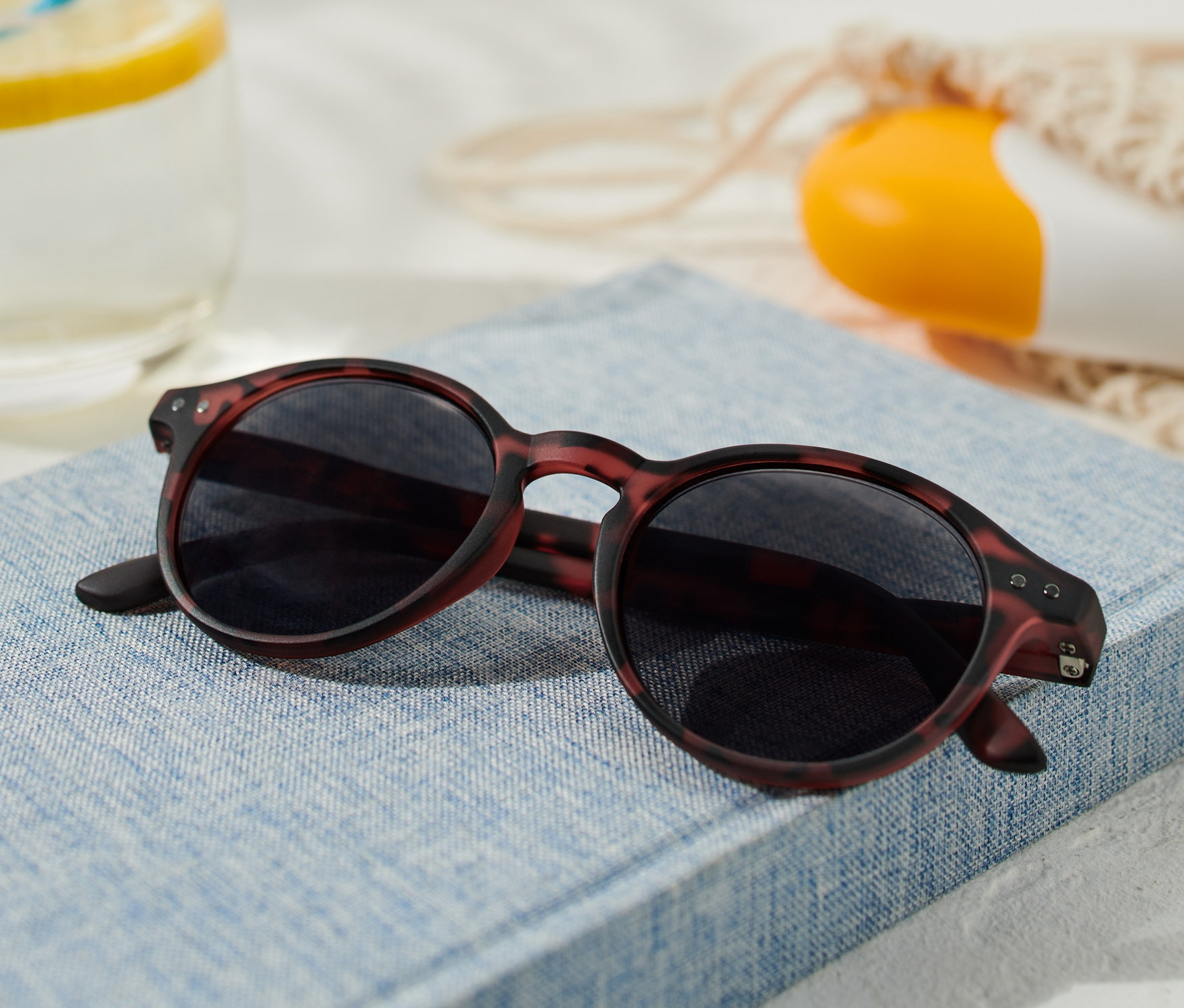 Egy napszemüveg egy kék könyvön fekszik, mellette egy pohár citrommal és egy naptej.