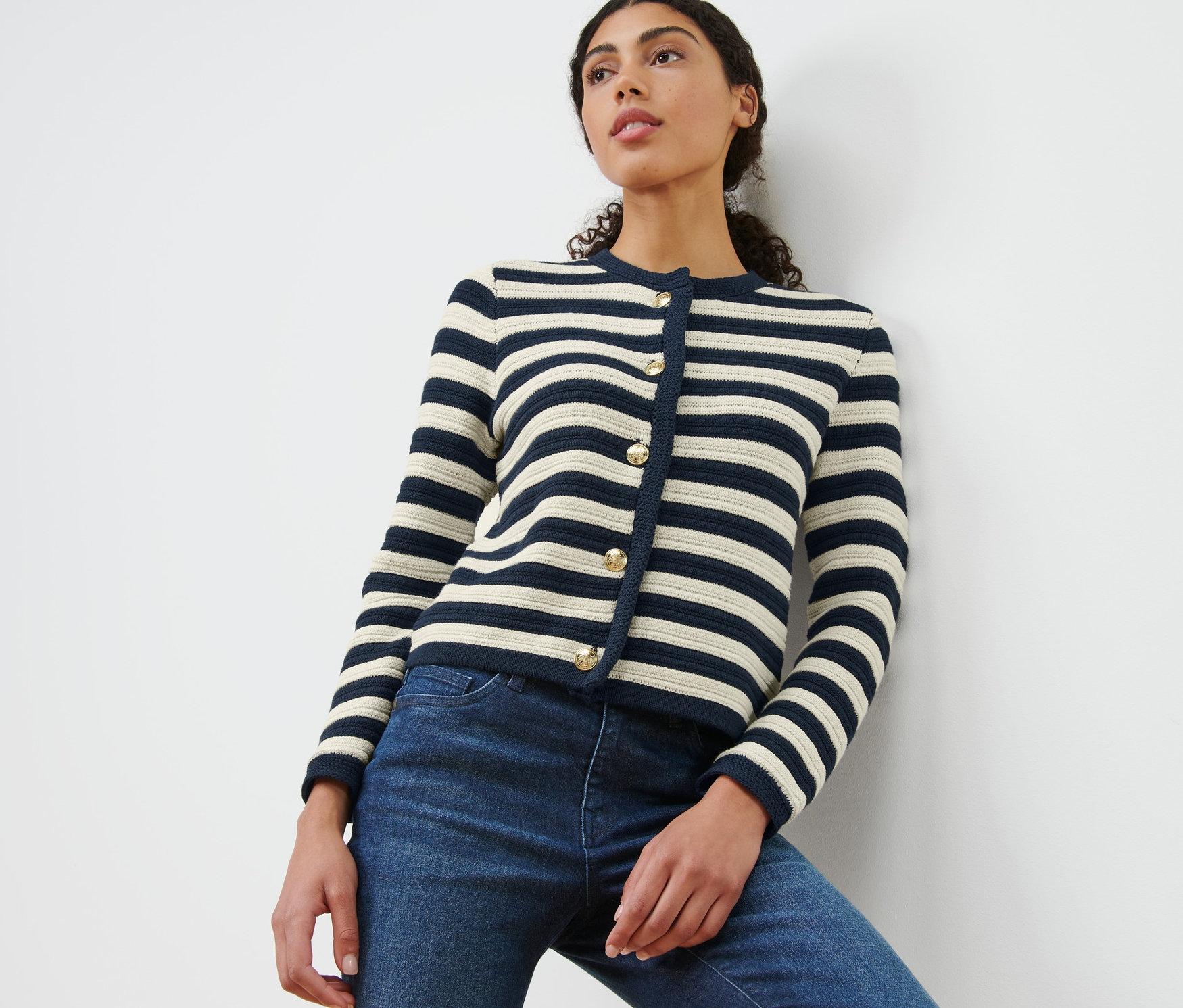 Egy fehér falnak támaszkodó nő tengerészkék és krémszínű csíkos, strukturált kötésű kardigánt és farmert visel.