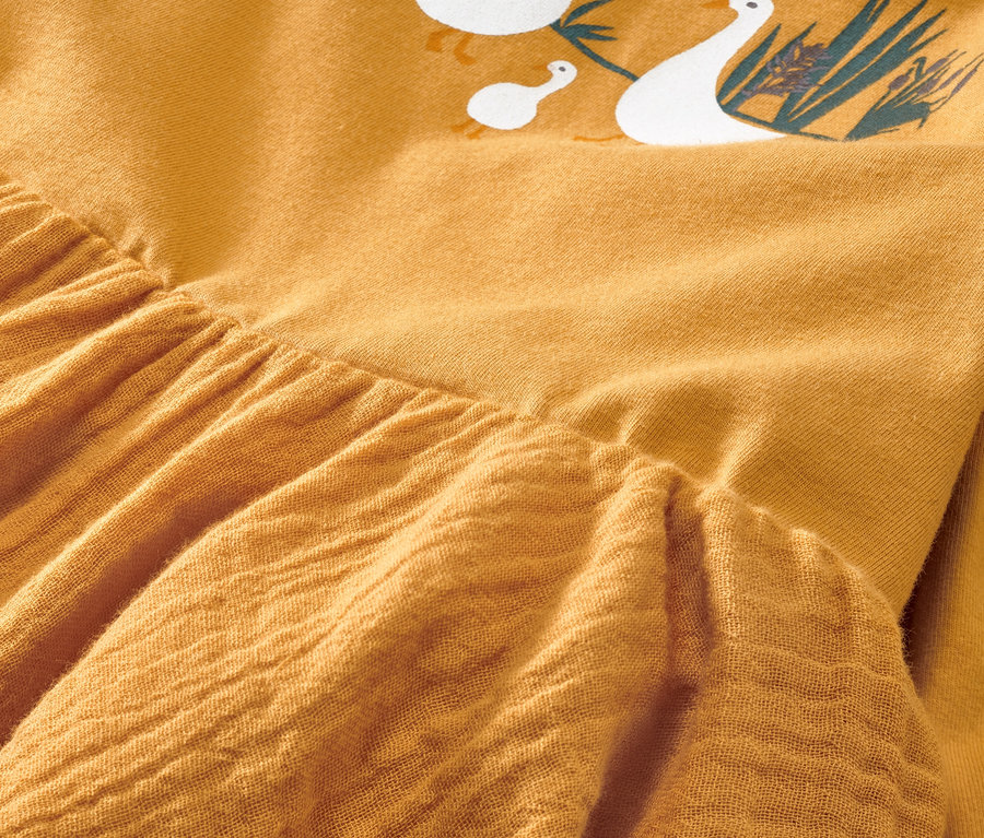 Egy sárga, szövött baba ruha részletének közeli képe, liba mintával.