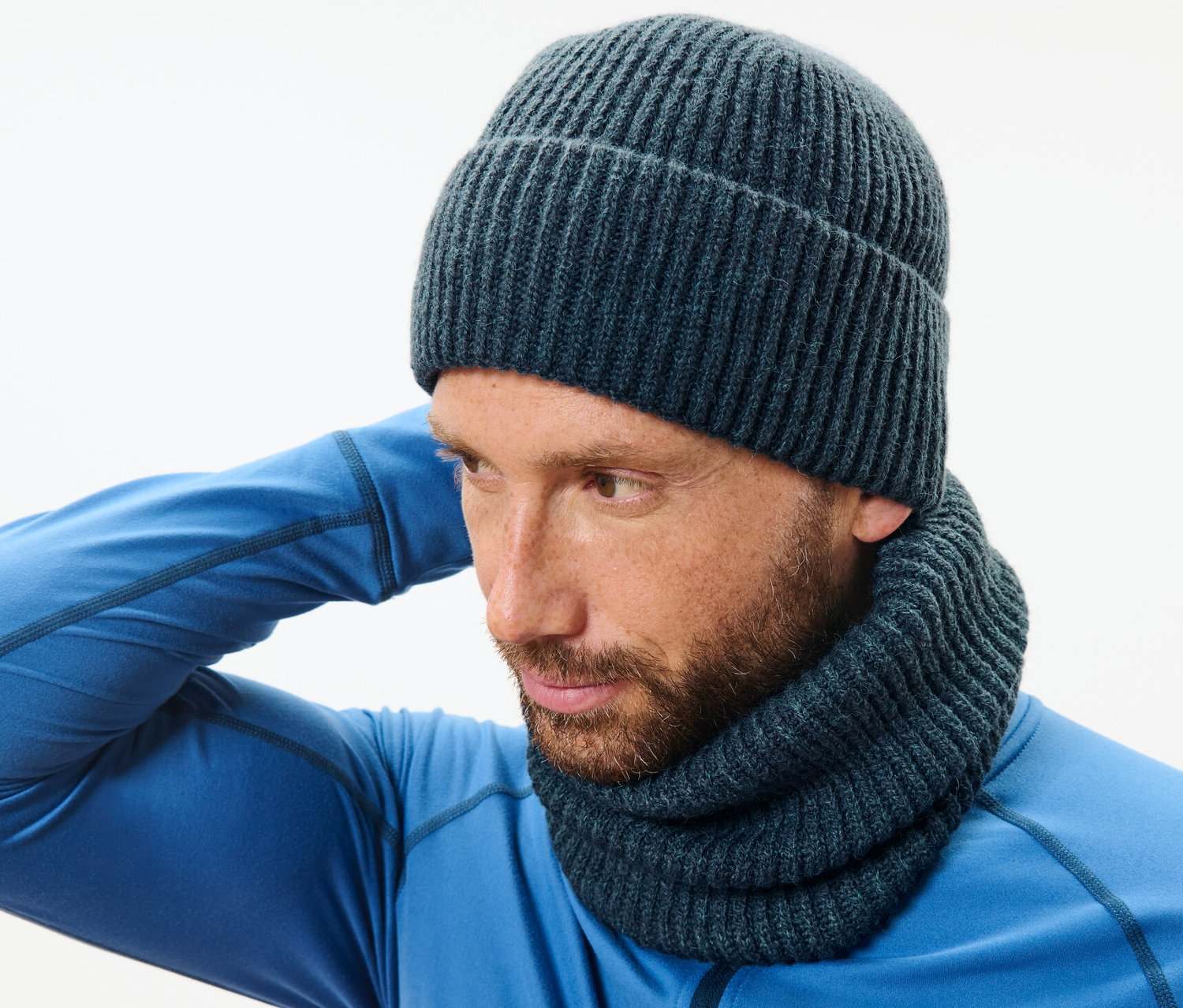 Kék pulóvert és sötétkék, polárbéléssel ellátott kötött csősálat viselő férfi igazgatja a kötött sapkáját.