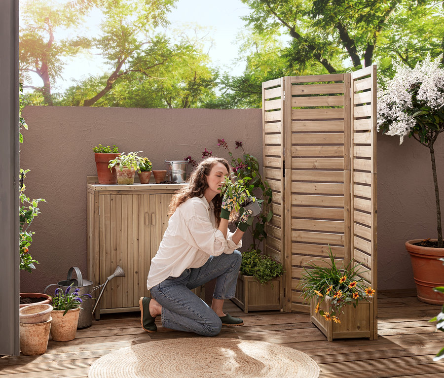 Egy nő térdel egy teraszon, és egy növényt szagol a »Tinus« flexibilis paraván növényládákkal és a »Tinus« kis kerti tároló közelében.