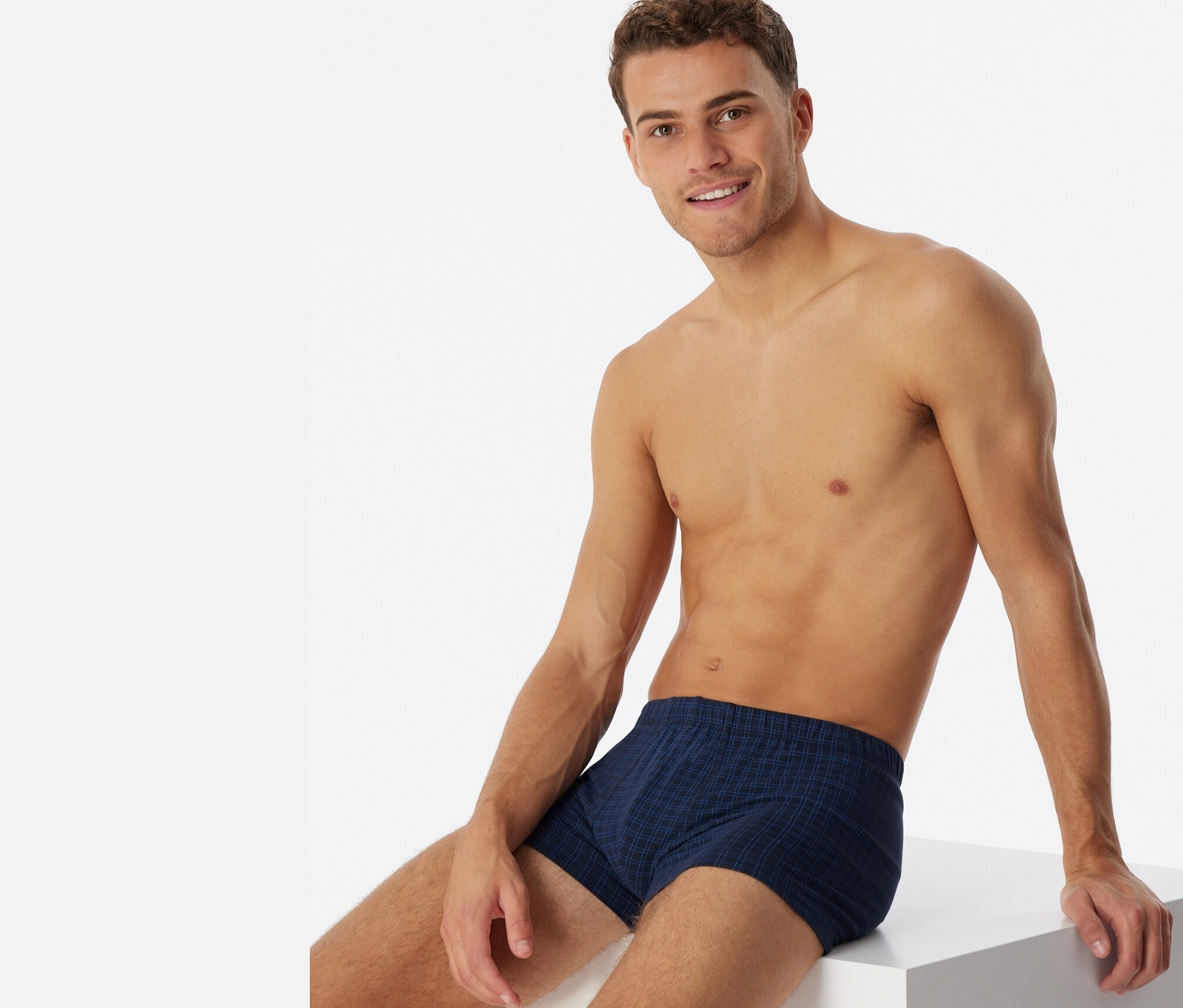 Egy férfi ül egy fehér kockán, kockás SCHIESSER sortot visel.