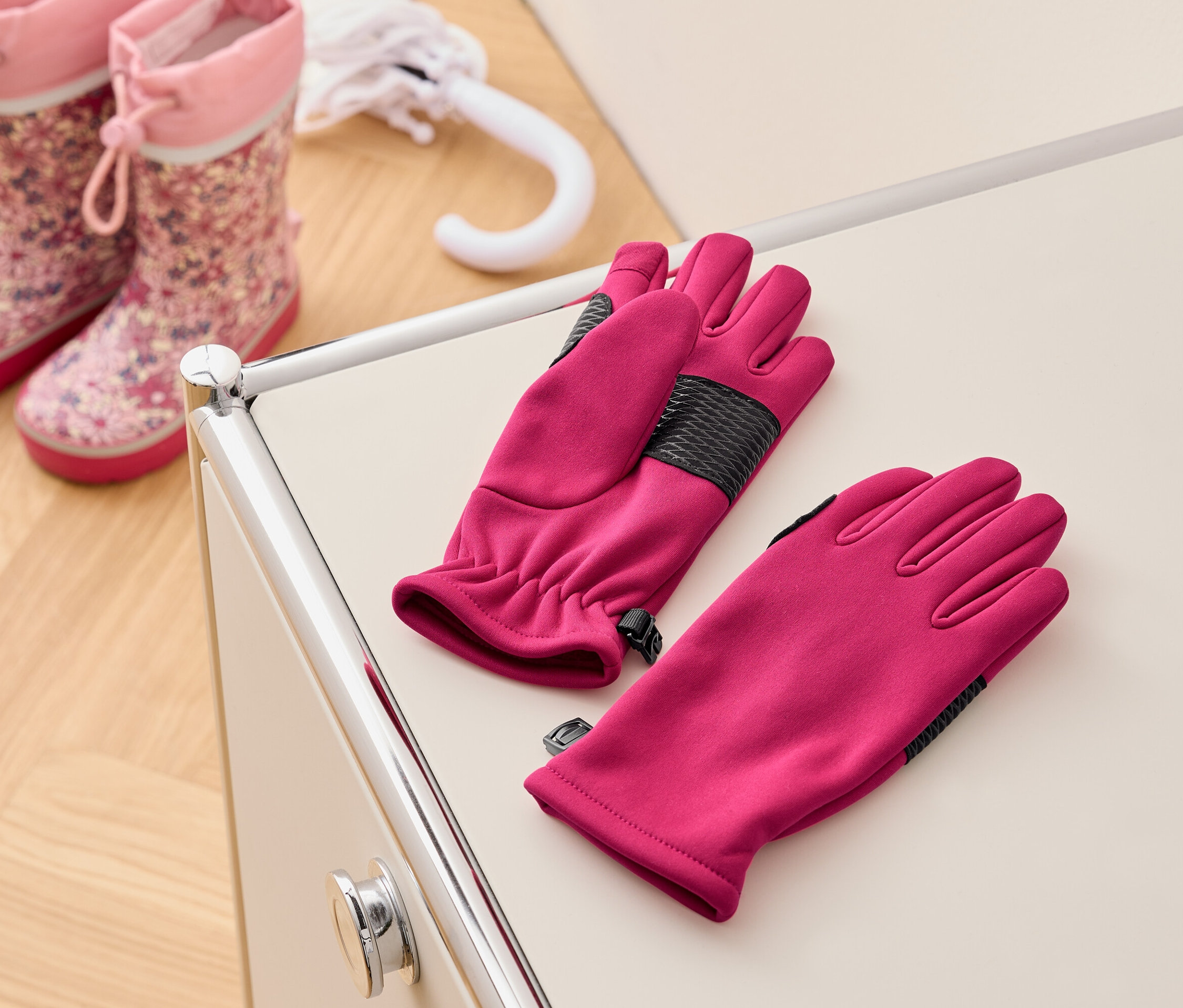 Egy pár rózsaszín kesztyű egy krémszínű asztalon fekszik rózsaszín gyerek gumicsizmák és egy fehér esernyő mellett.