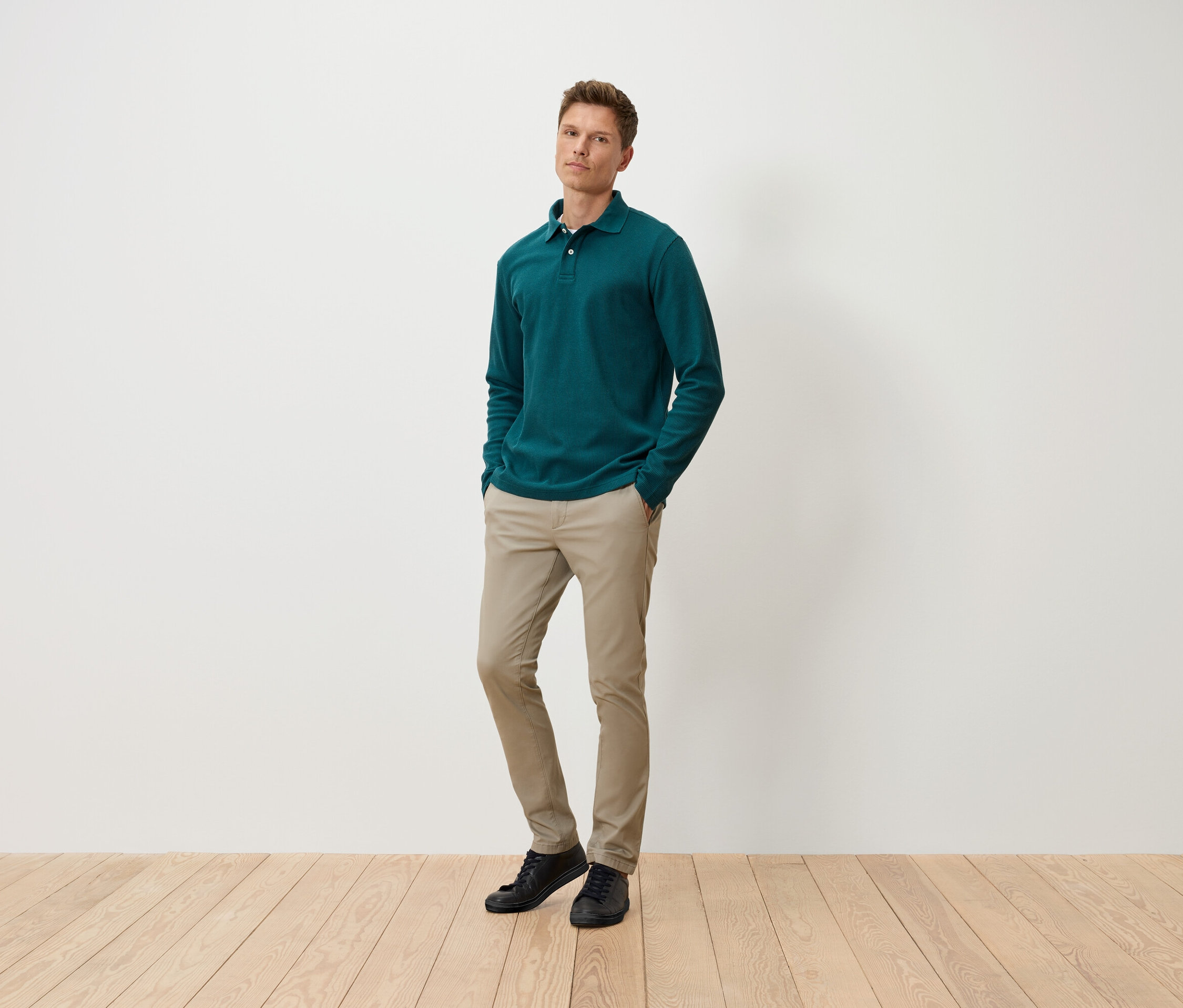 Egy férfi áll zsebre tett kézzel, zöld hosszú ujjú pólóban, bézs nadrágban és fekete cipőben.