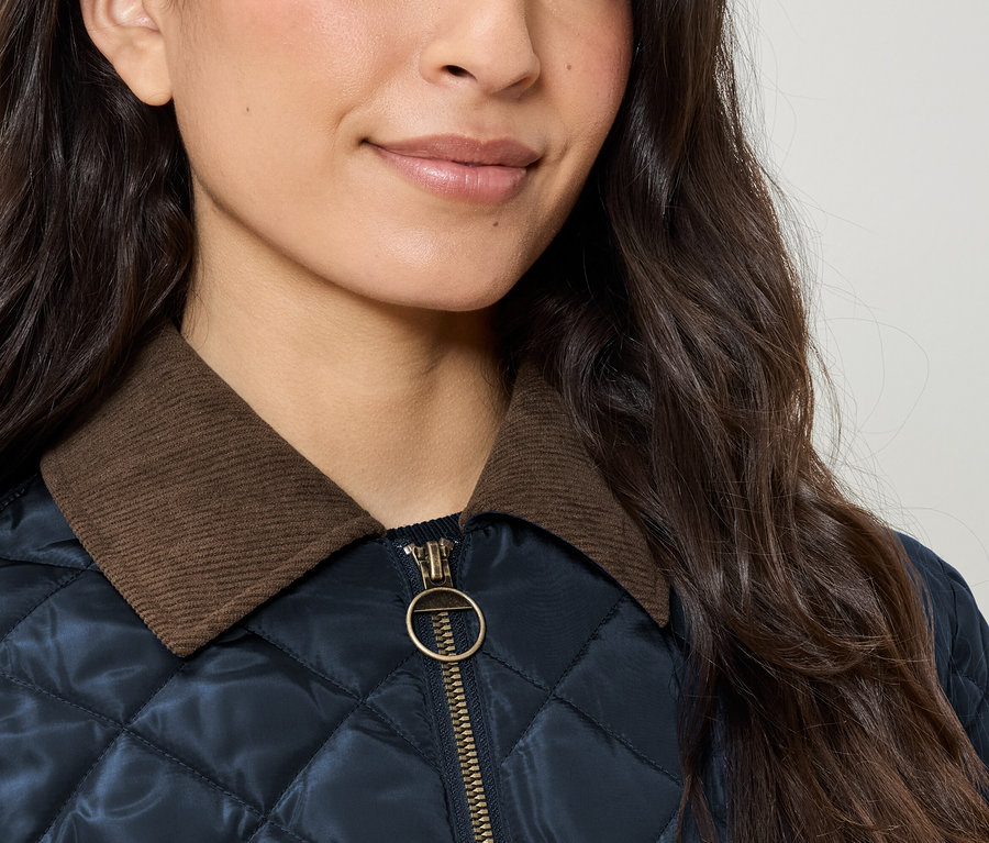 Egy női steppelt dzseki részlete barna kordbársony gallérral és arany cipzárral.