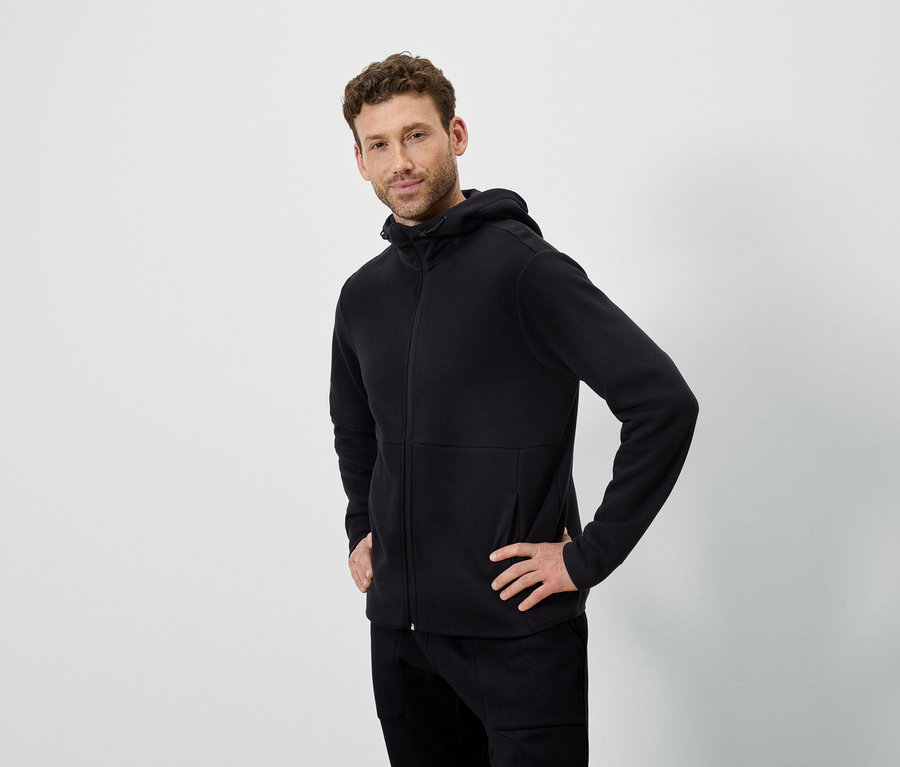 Egy férfi pózol fehér háttér előtt. Fekete kapucnis pulóvert és fekete nadrágot visel.