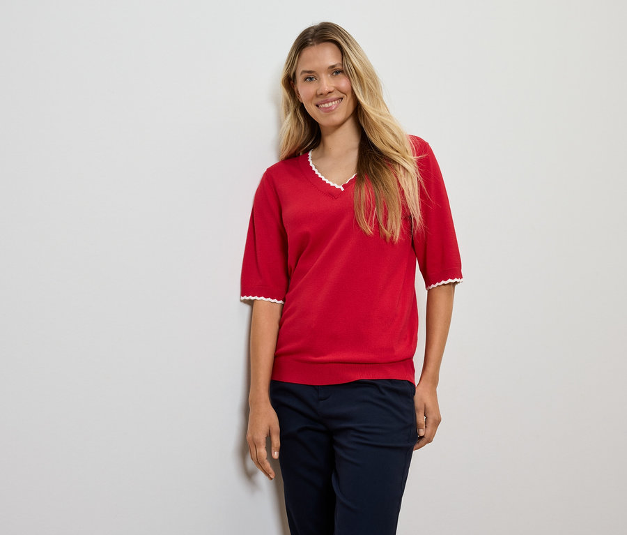 Egy szőke hajú nő piros pulóverben és sötétkék nadrágban pózol.