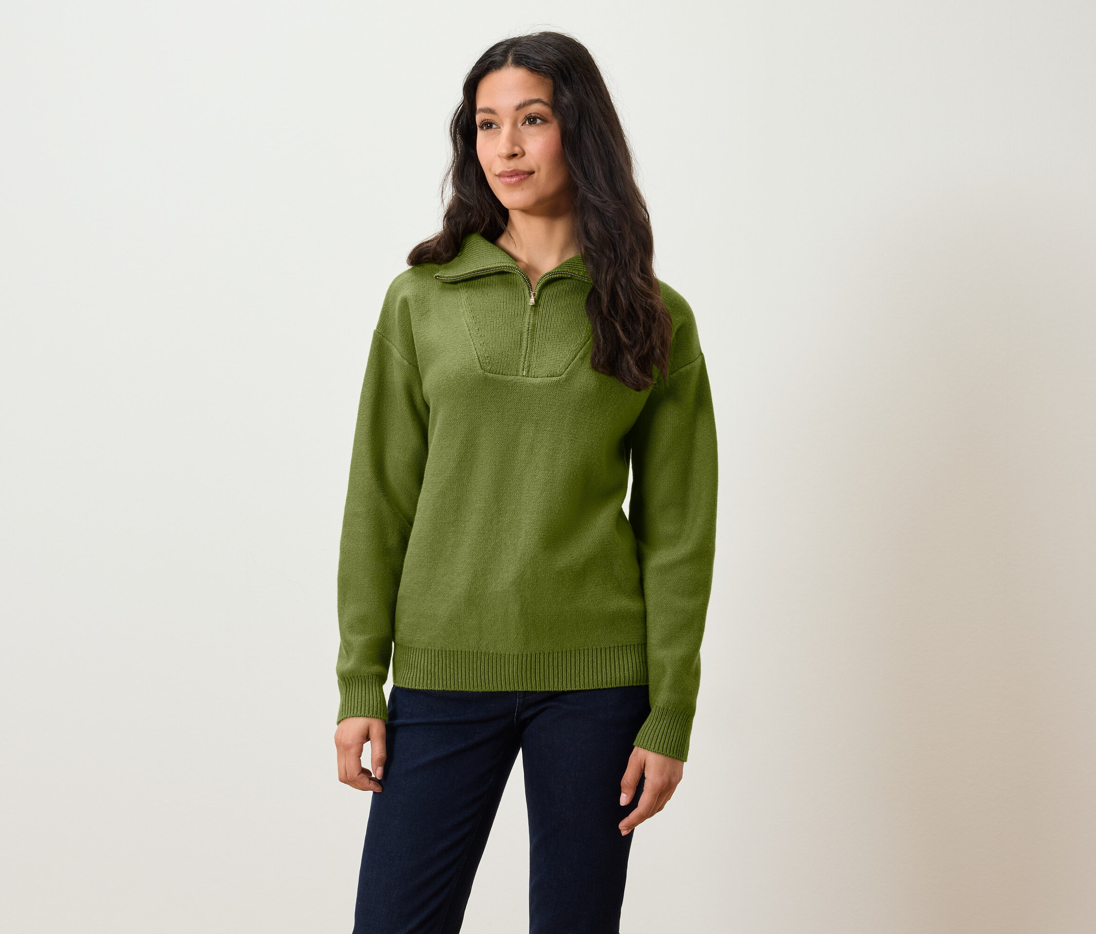 Egy nő áll, olívazöld, rövid cipzáros pulóvert és sötétkék nadrágot visel.