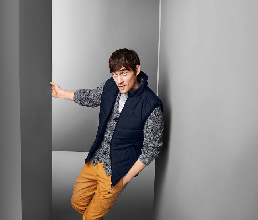 Férfi a falnak dőlve, steppelt mellényt, pulóvert és nadrágot visel.