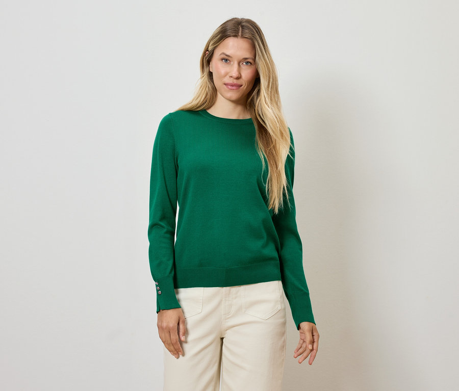 Szőke nő zöld pulóverben és krémszínű nadrágban áll fehér háttér előtt.