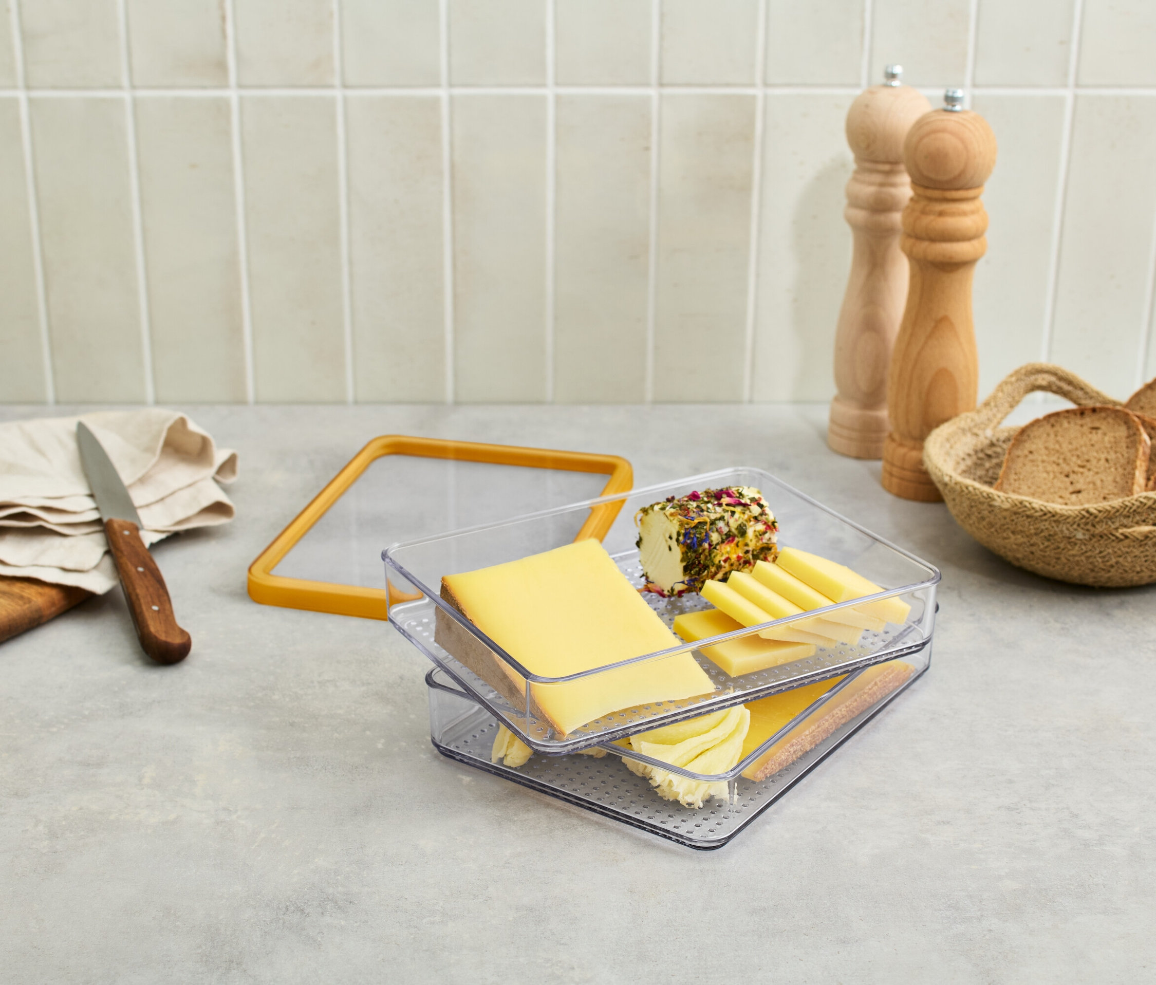 Az asztalon sajt van egy átlátszó edényben, szőlő, kés, kenyér egy kosárban és fűszermalom.