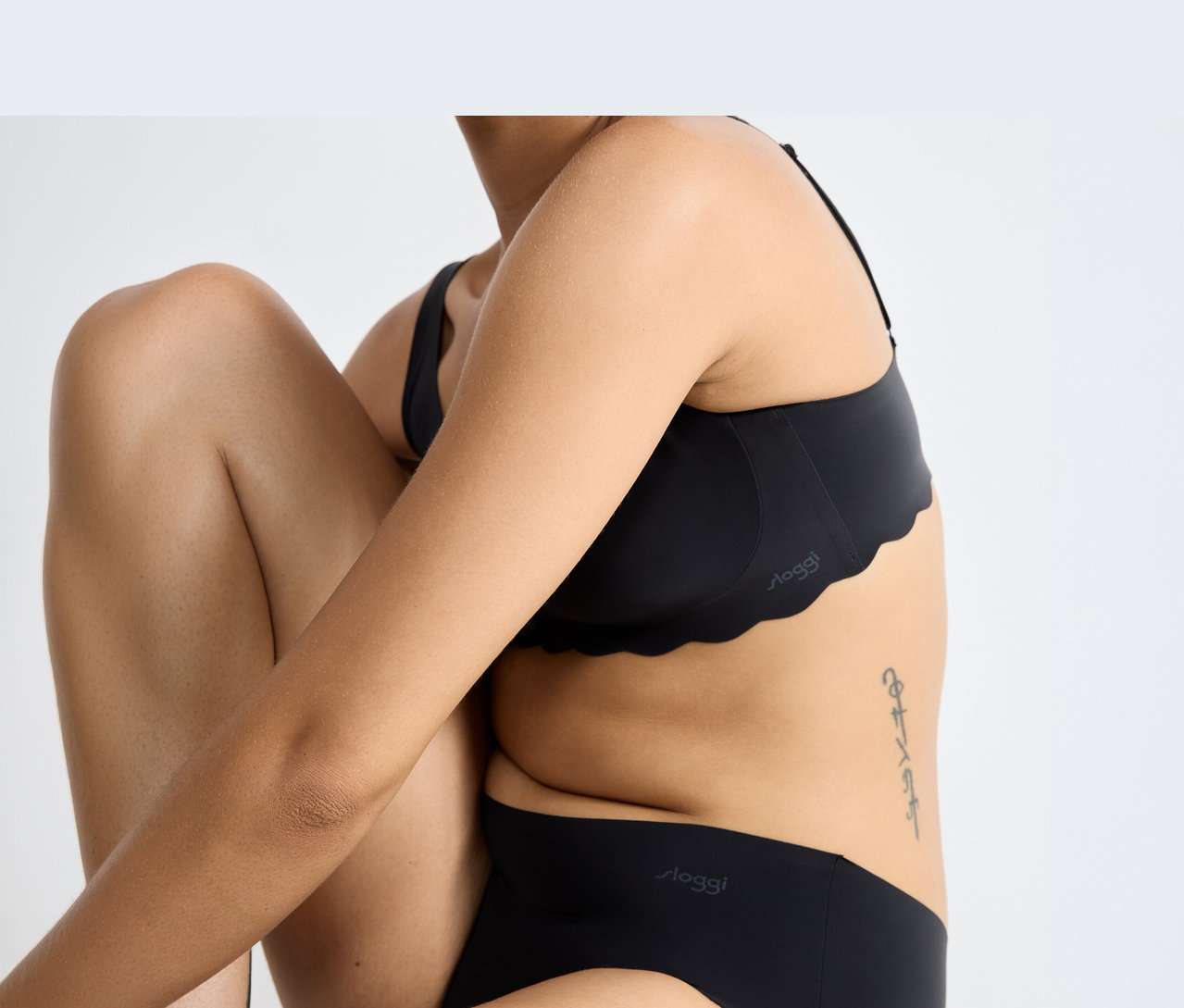 Egy személy fekete sloggi ZERO Microfibre 2.0 Soft melltartót visel hullámos széllel és hozzá illő fekete sloggi alsóneműt, behajlított térdekkel és keresztezett karokkal ülve. Az oldalán tetoválás látható.