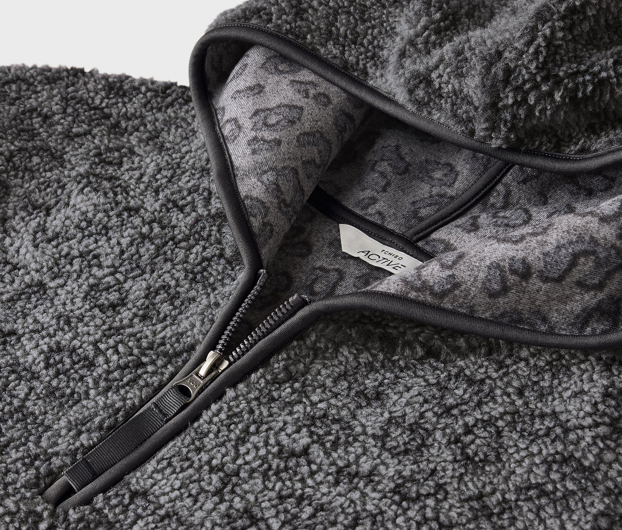 Egy szürke, bolyhos kapucnis pulóver részlete cipzárral és szürke állatmintával a kapucni belső oldalán.