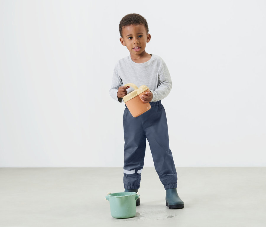 Egy kisfiú narancssárga vödröt tart a kezében, kék esőnadrágot és gumicsizmát visel.