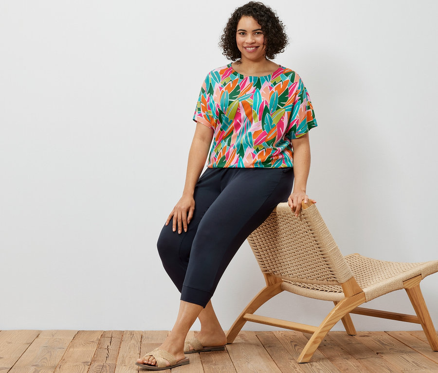 Egy nő egy fonott széken ül, sötétkék 3/4-es jersey nadrágot és nyomott mintás, ejtett vállú pólóblúzt visel.